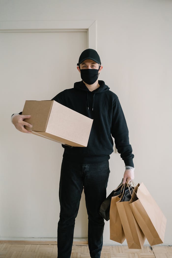 A delivery man wearing a mask holds a box and shopping bags indoors.
